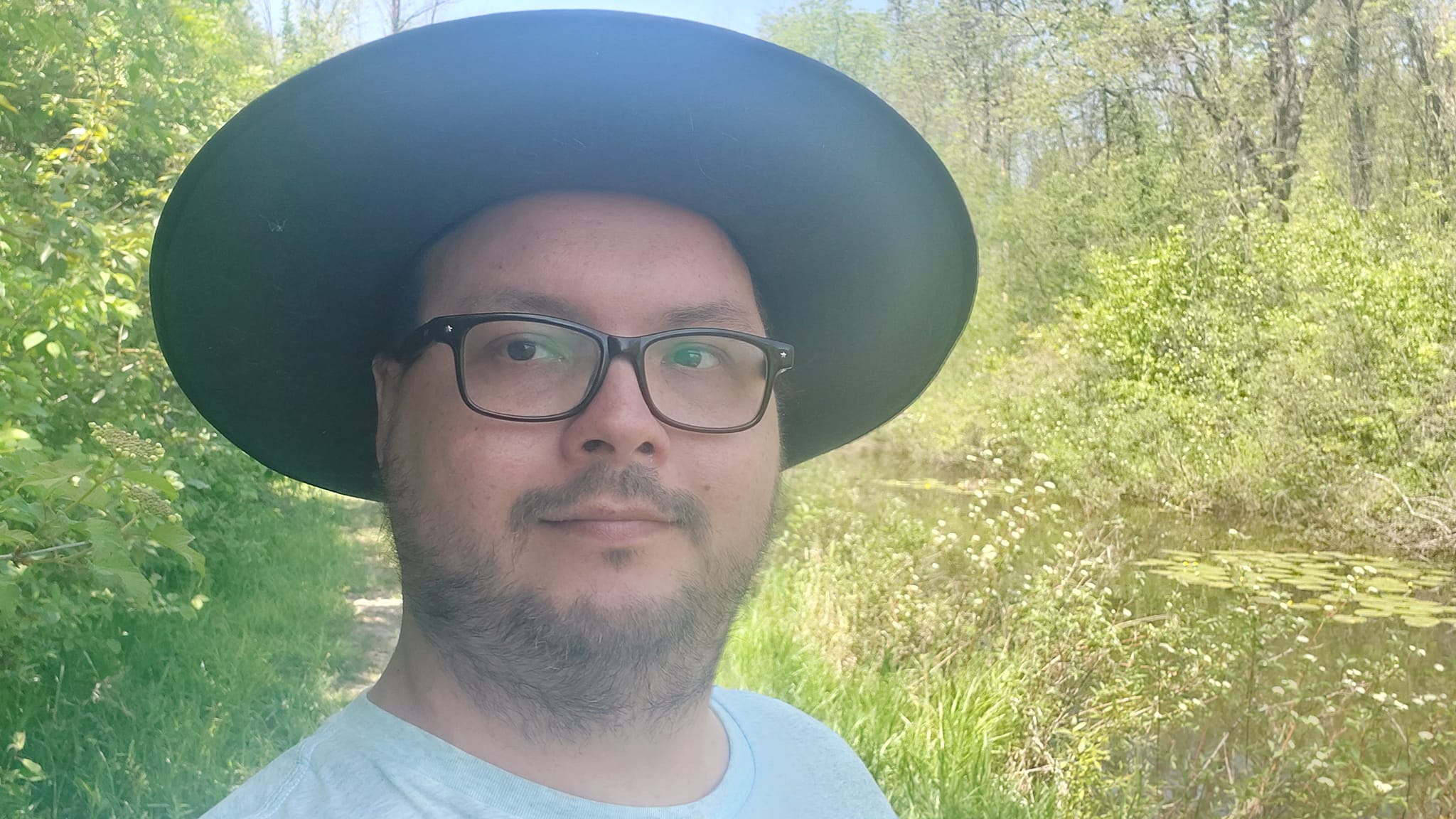 A man wearing a blue shirt wears a wide brimmed sunhat in a forest next to a ditch